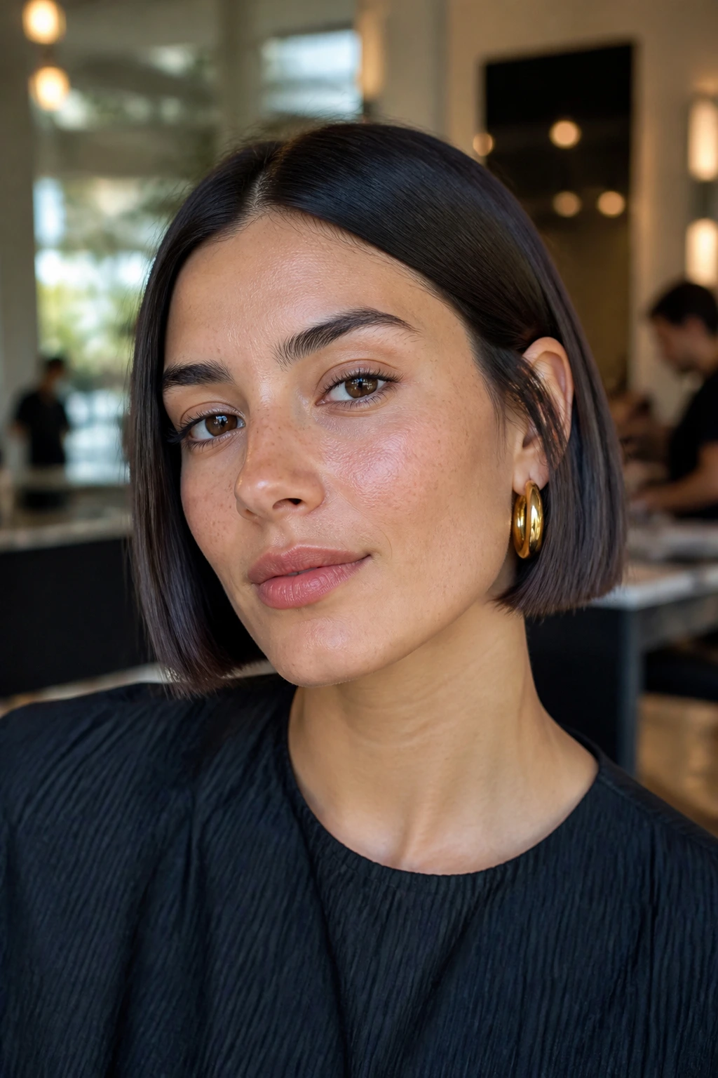 Close-up portrait of a real woman with blunt no-bang French bob under warm salon lighting.