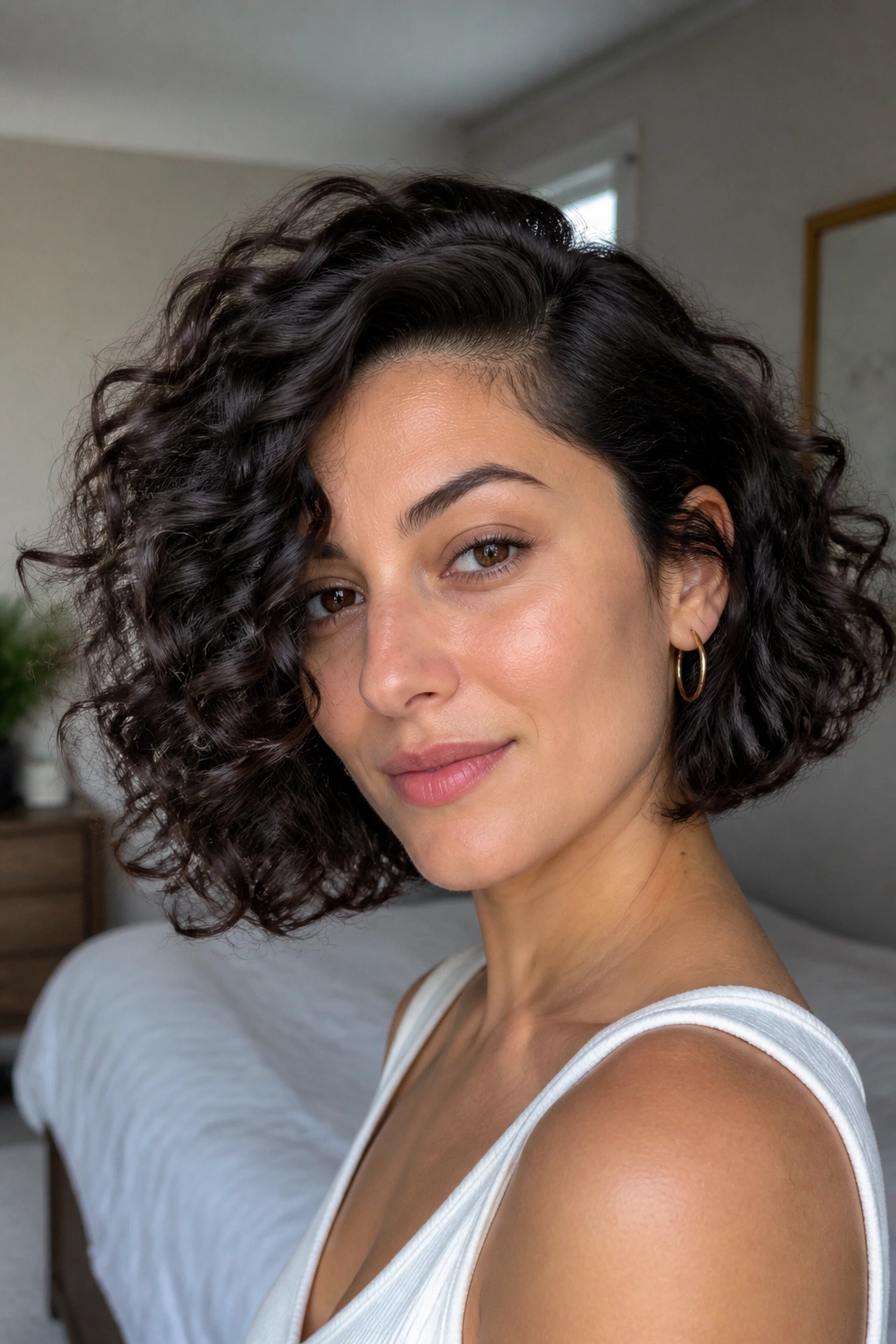 Close-up portrait of a woman with a deep side-part curly bob in a softly lit bedroom.